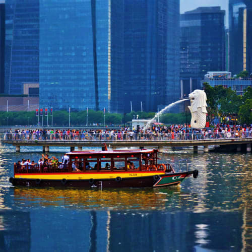 Red and yellow river boat cruising past the Merlion statue with modern skyscrapers and crowds along Singapore's waterfront