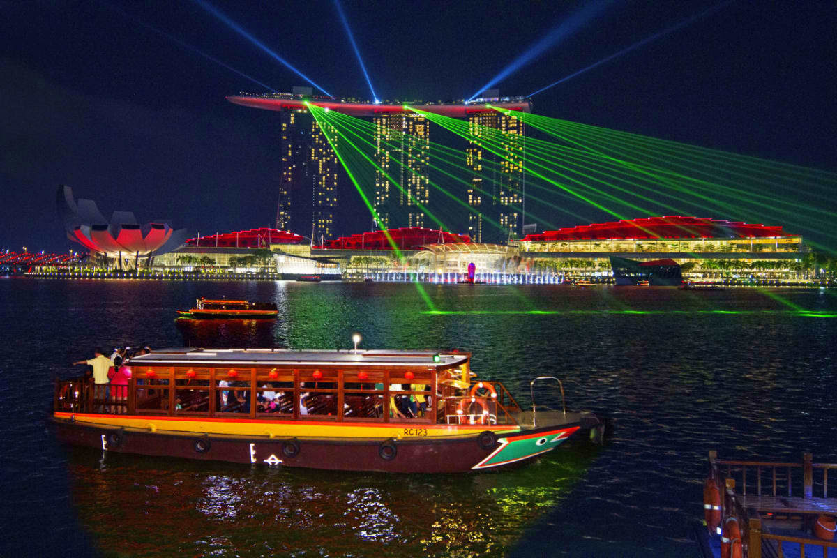 Traditional wooden river boat with tourists cruising past illuminated Marina Bay Sands hotel with laser light show at night i