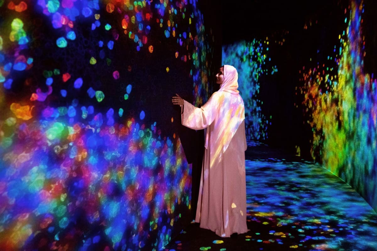 TeamLab Phenomena Person in white clothing standing in immersive digital art installation with colorful projected lights and particles on walls