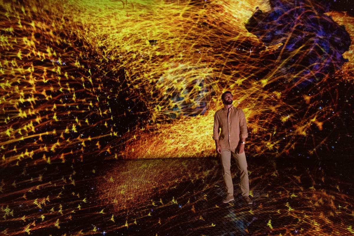 TeamLab Phenomena Man standing in immersive digital art installation with glowing golden and orange light projections creating abstract pattern