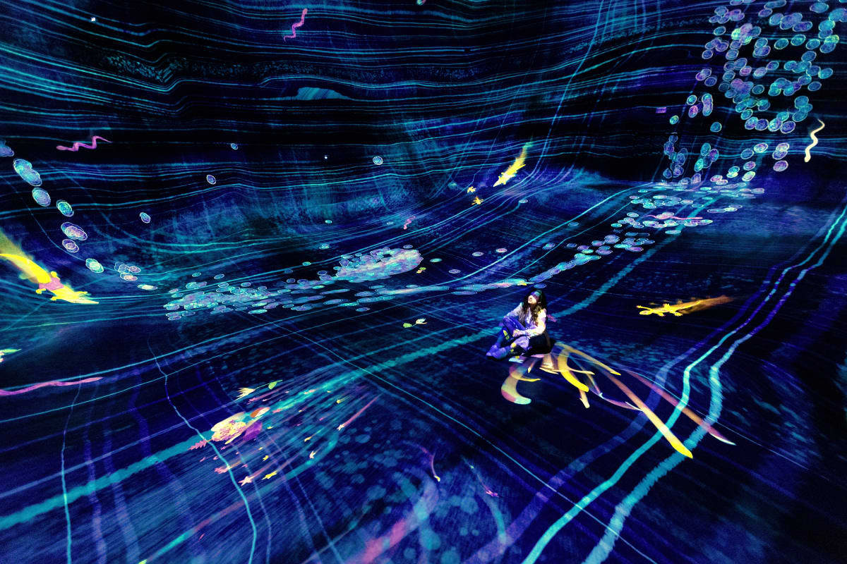 TeamLab Phenomena Person sitting in immersive digital art installation with flowing cyan and yellow light streams and glowing spheres in dark s