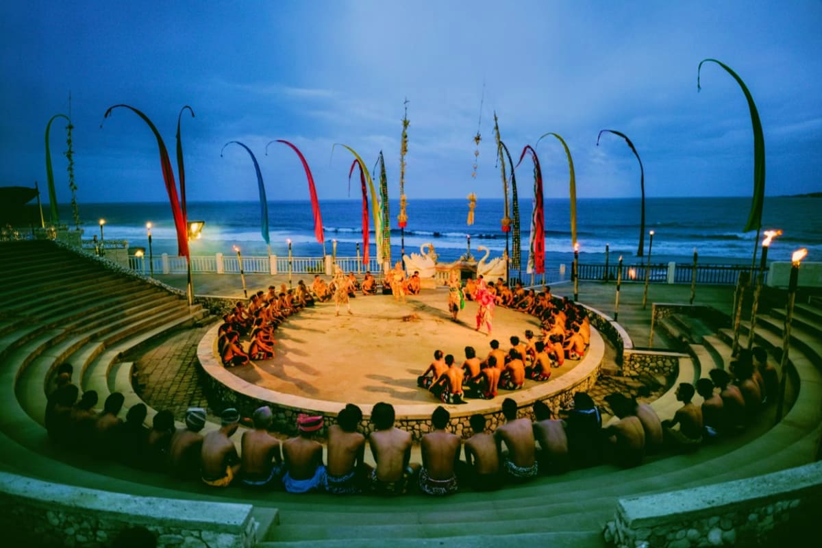 Balinese performers in orange sarongs dancing in circular amphitheater at Uluwatu Temple with ocean and colorful flags in bac