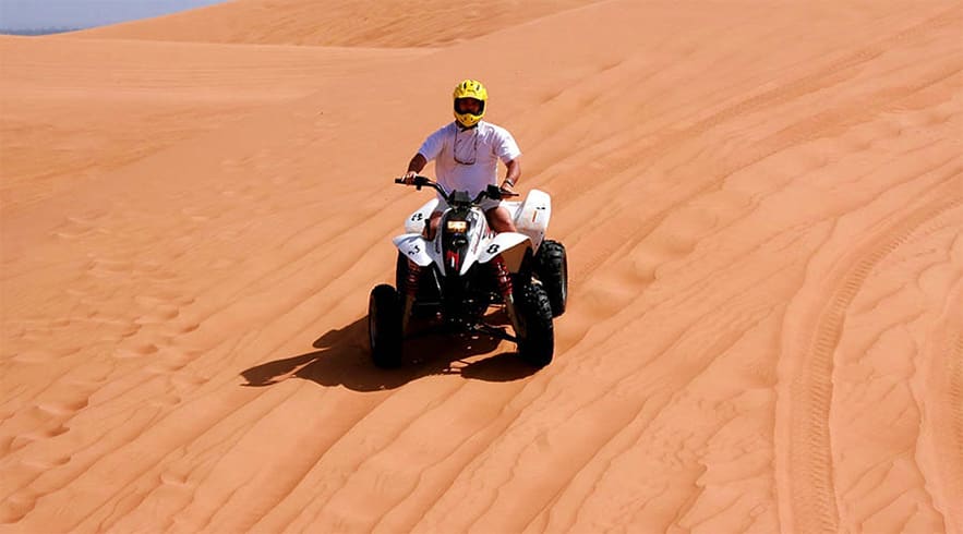 Person riding white and black quad bike across sandy desert dunes in Abu Dhabi, wearing yellow helmet and white shirt