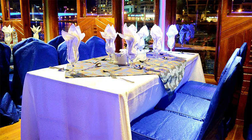 Elegant dining table setup on a dhow cruise with white linens, blue chairs, and city lights visible through large windows at