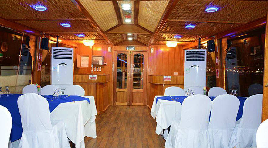 Interior of traditional wooden dhow cruise vessel with dining tables set with white and blue linens, wooden ceiling and ambie