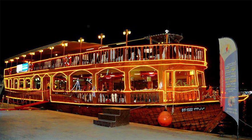 Dhow Cruise Abu Dhabi Illuminated traditional wooden dhow boat at night with multiple decks, ornate railings, and warm golden lighting for Abu Dhab
