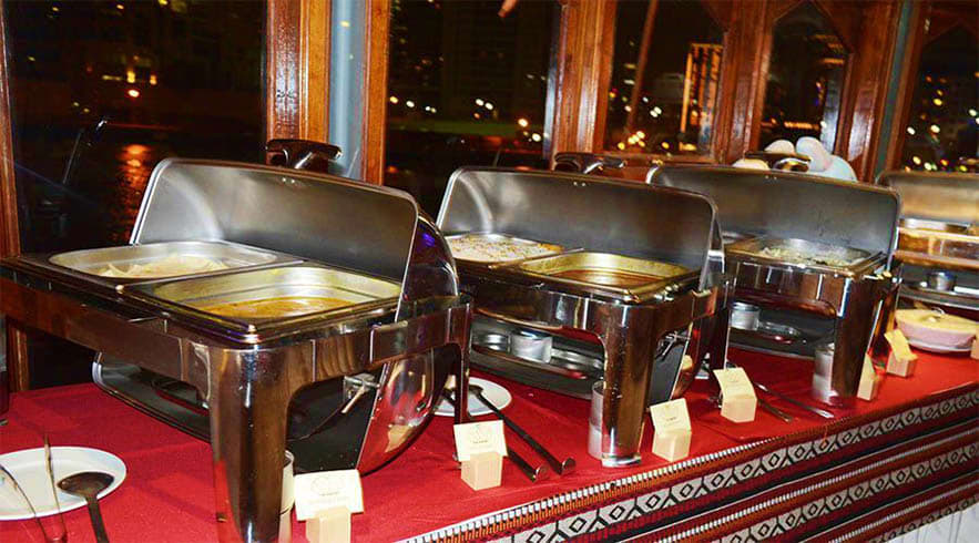 Dhow Cruise Abu Dhabi Three stainless steel chafing dishes with food warming lids on red carpet in elegant dining room with night city lights visib