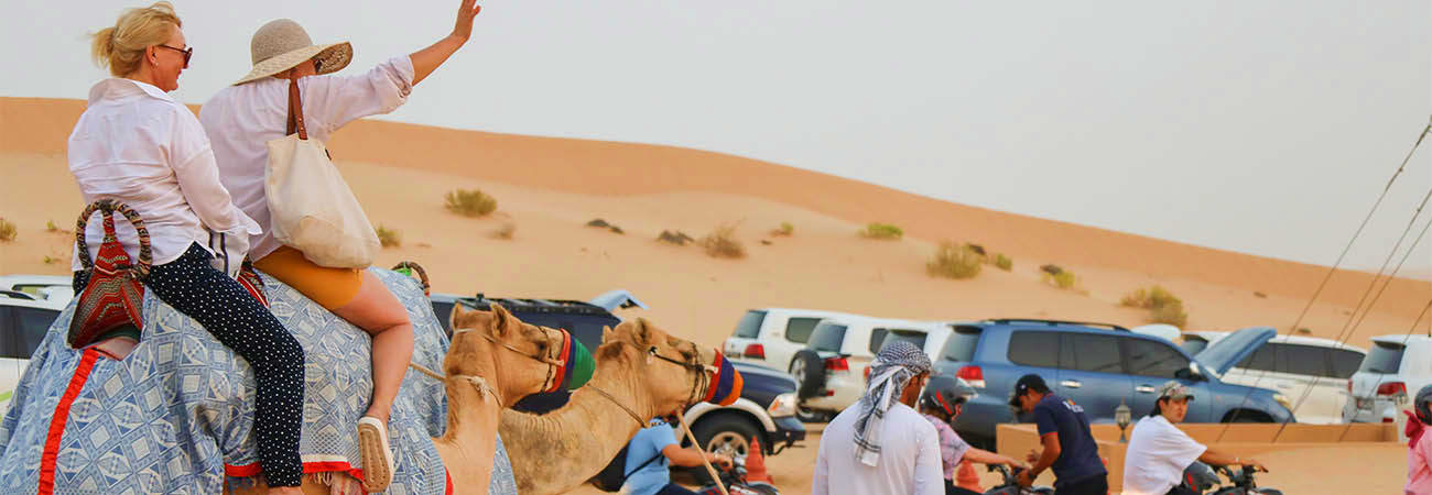 Sunrise Desert Safari Abu Dhabi Tourists riding camels in desert with vehicles and guides in background during Abu Dhabi desert safari tour