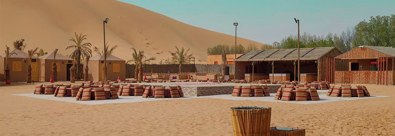 Sunrise Desert Safari Abu Dhabi Desert safari camp in Abu Dhabi with traditional structures, palm trees, and striped seating arrangements on sand dunes