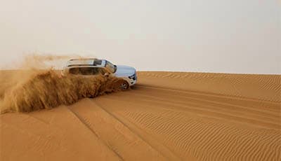Sunrise Desert Safari Abu Dhabi Silver SUV driving through sandy desert dunes creating dust clouds during desert safari in Abu Dhabi