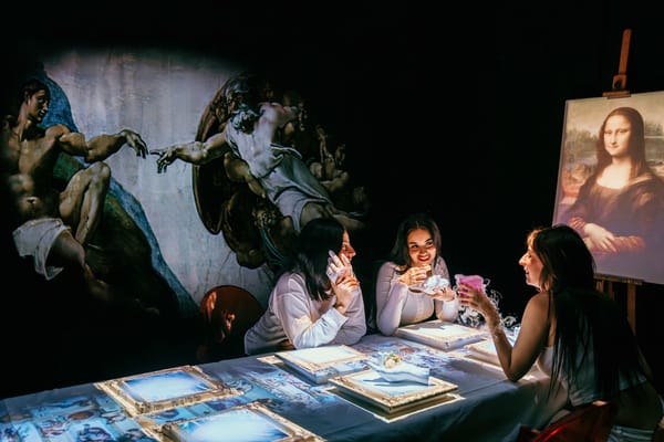 Seven Paintings Immersive Dining Experience Three women seated at a dining table with art books, surrounded by projected Renaissance paintings including the Creation of