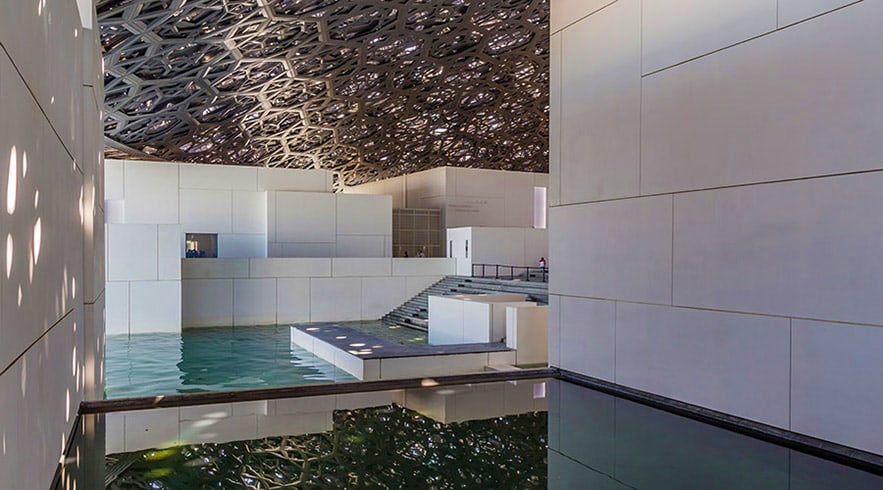 Interior view of Louvre Abu Dhabi showing modern white architecture, geometric ceiling design, and reflecting pool with water