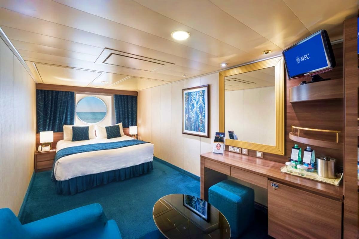MSC Splendida South America Journey MSC cruise ship cabin with queen bed, blue carpet, wood furnishings, porthole window, TV, and seating area