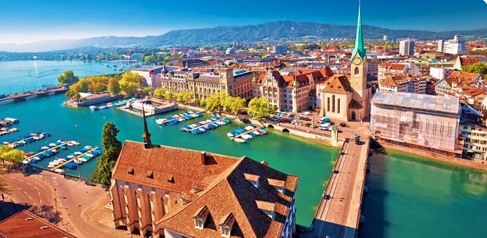 Swiss Capitals Aerial view of Zurich's old town with Church of St. Peter, turquoise Limmat River, boats, and Alpine mountains in background