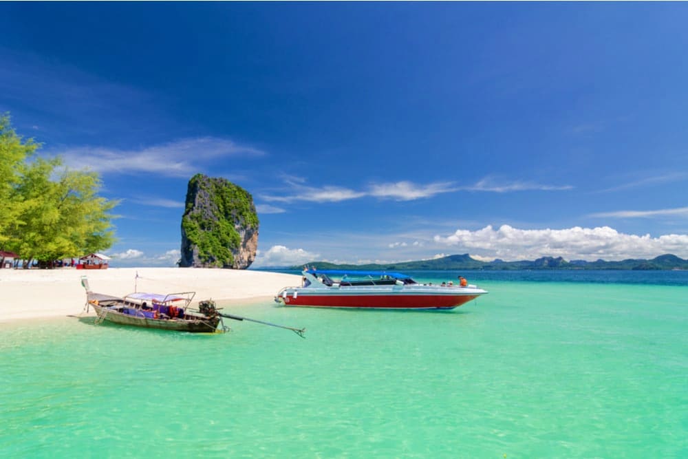 Tropical beach scene in Phuket Krabi with turquoise water, white sand, traditional long-tail boats, and a limestone rock form