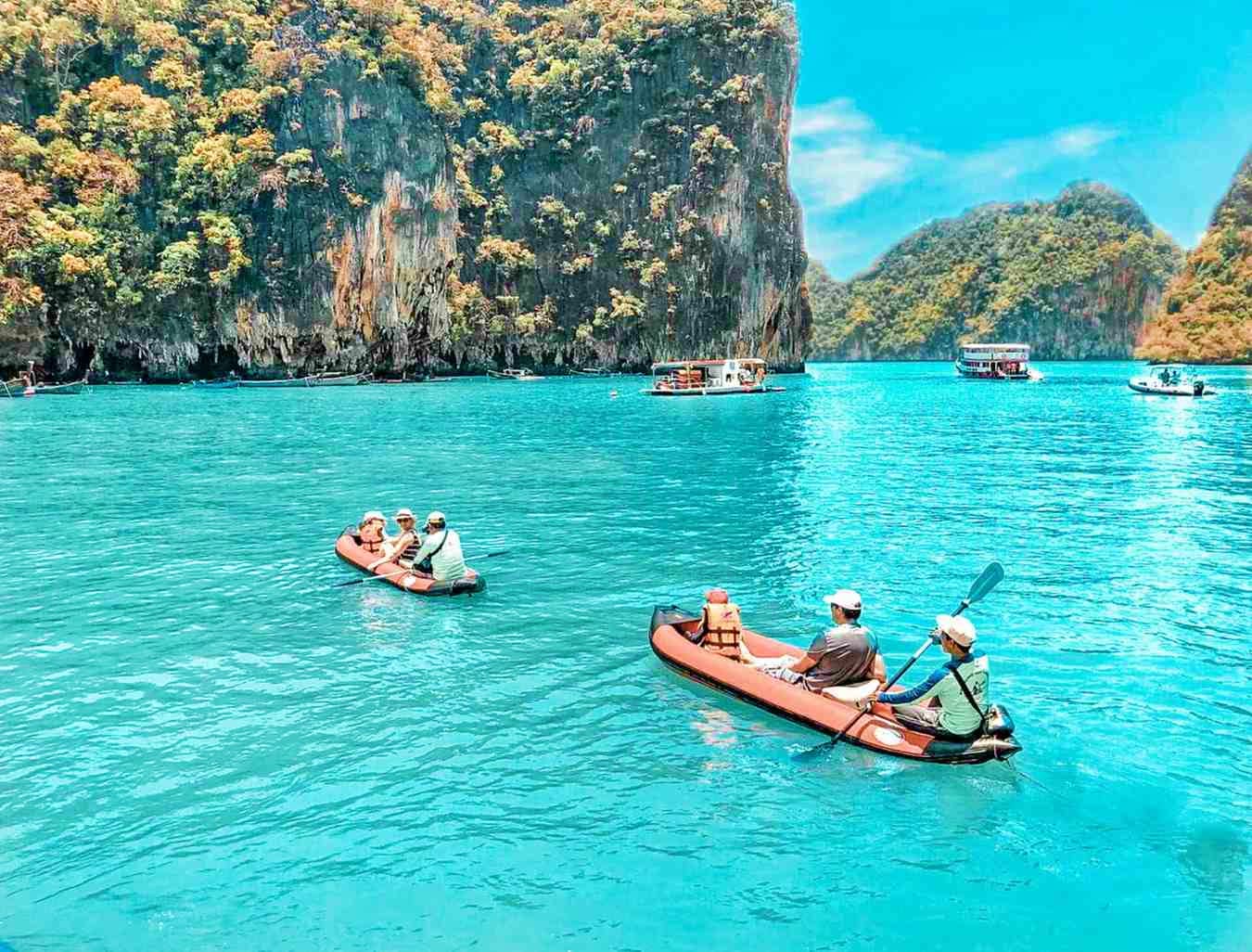 Tourists kayaking in turquoise water near dramatic limestone cliffs in Phang Nga Bay, Phuket, Thailand with boats in backgrou