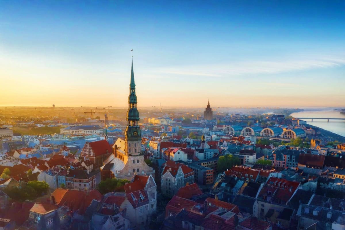 Aerial sunrise view of Riga's Old Town with St. Peter's Church spire, red-roofed buildings, and Daugava River in background