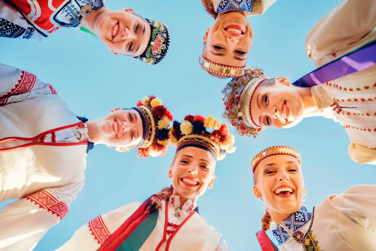 Happy children wearing traditional Latvian folk costumes and embroidered clothing gathered in a circle against a blue sky