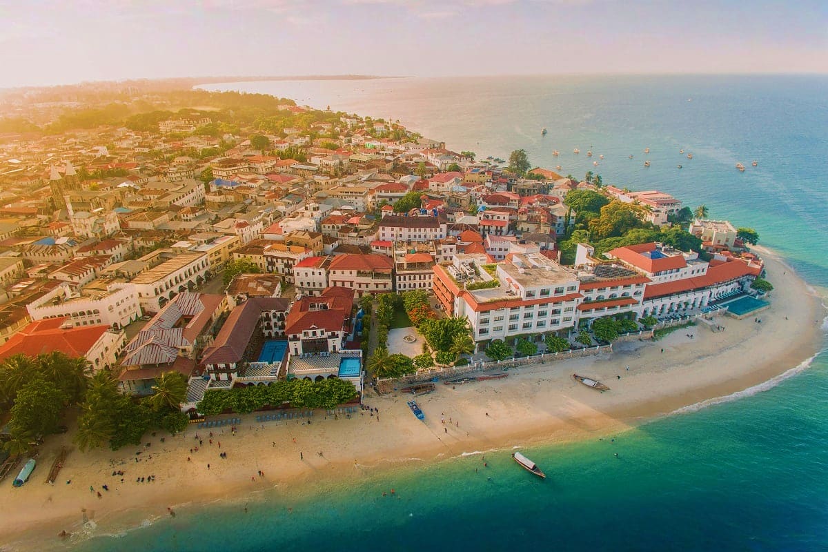 Aerial view of a coastal town with terracotta-roofed buildings, sandy beach, turquoise water, and boats moored offshore at su