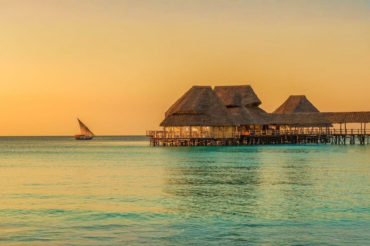 Overwater bungalow resort with thatched roofs at sunset in Zanzibar, with traditional sailing dhow in turquoise waters