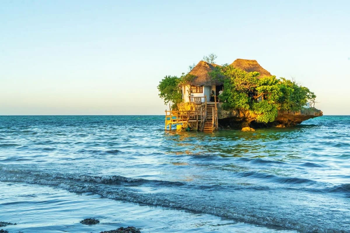 Thatched-roof stone house built on a rocky island surrounded by turquoise ocean water with yellow dock and wooden stairs, Zan