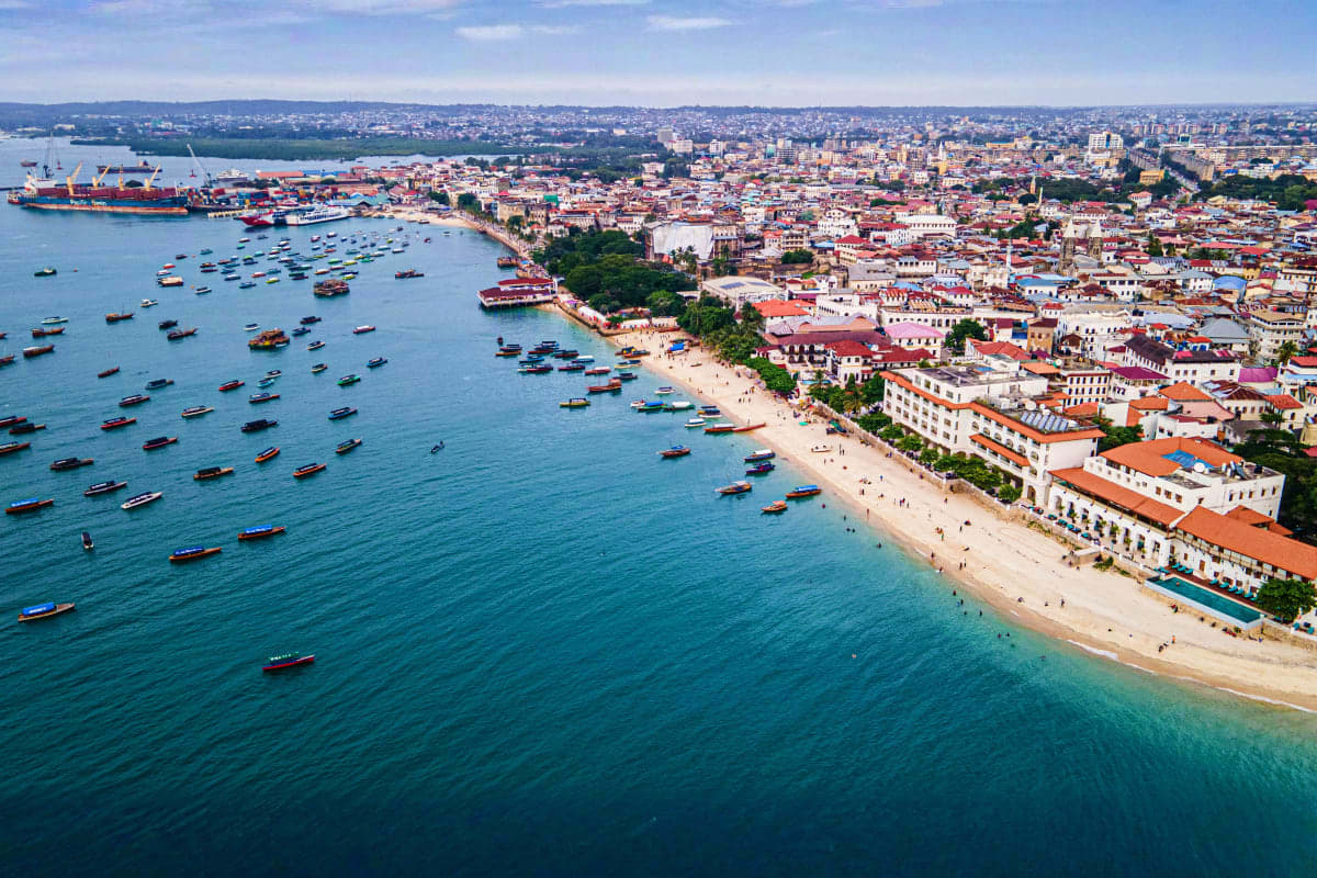 Aerial view of Zanzibar's Stone Town beachfront with turquoise water, colorful fishing boats, sandy beach, and historic coral