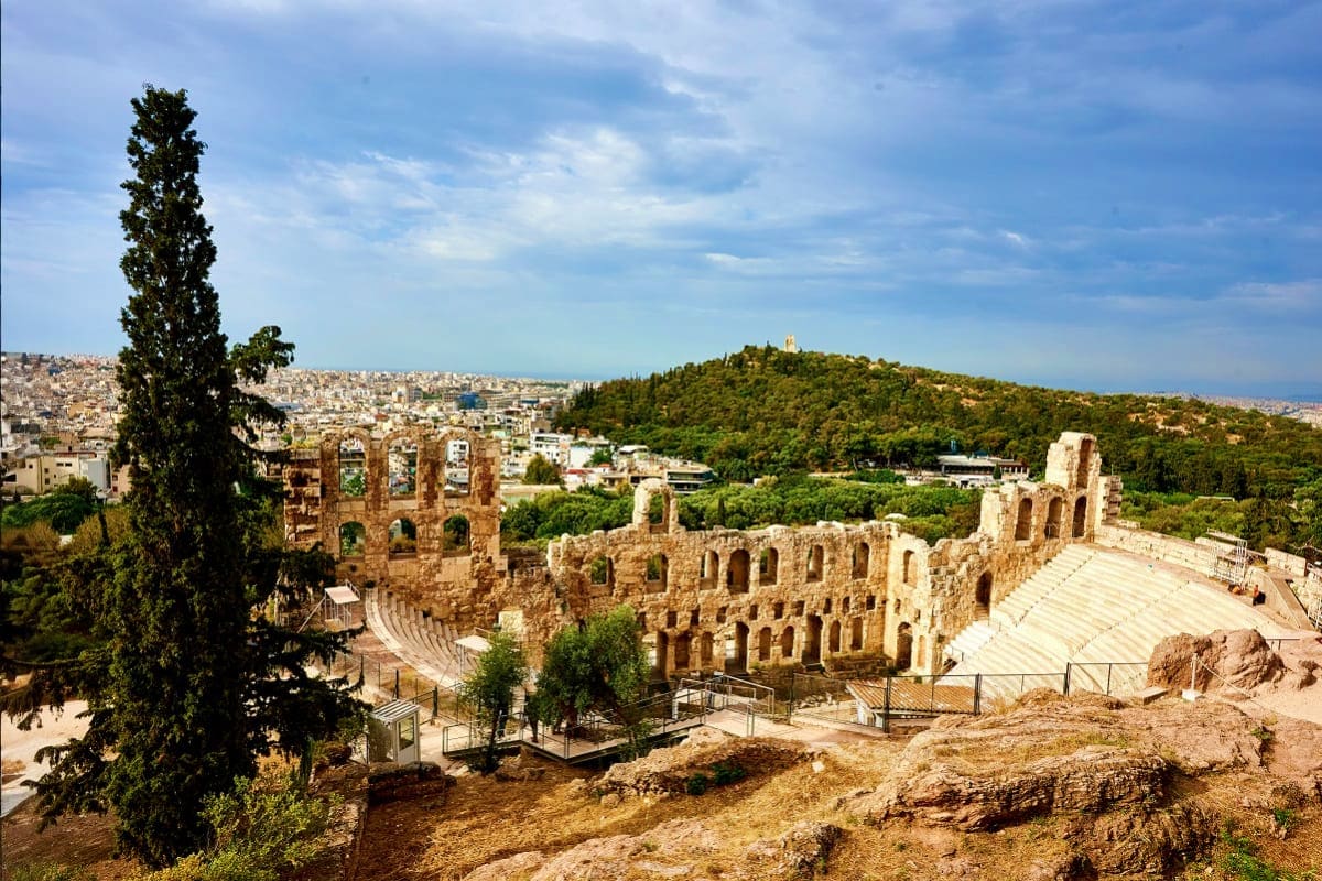 Ancient stone theater ruins with semicircular seating overlooking Athens cityscape and forested hills under blue sky