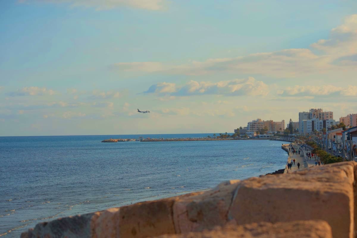 Mediterranean coastal town with beach promenade, modern buildings, calm turquoise sea, and helicopter flying overhead under c