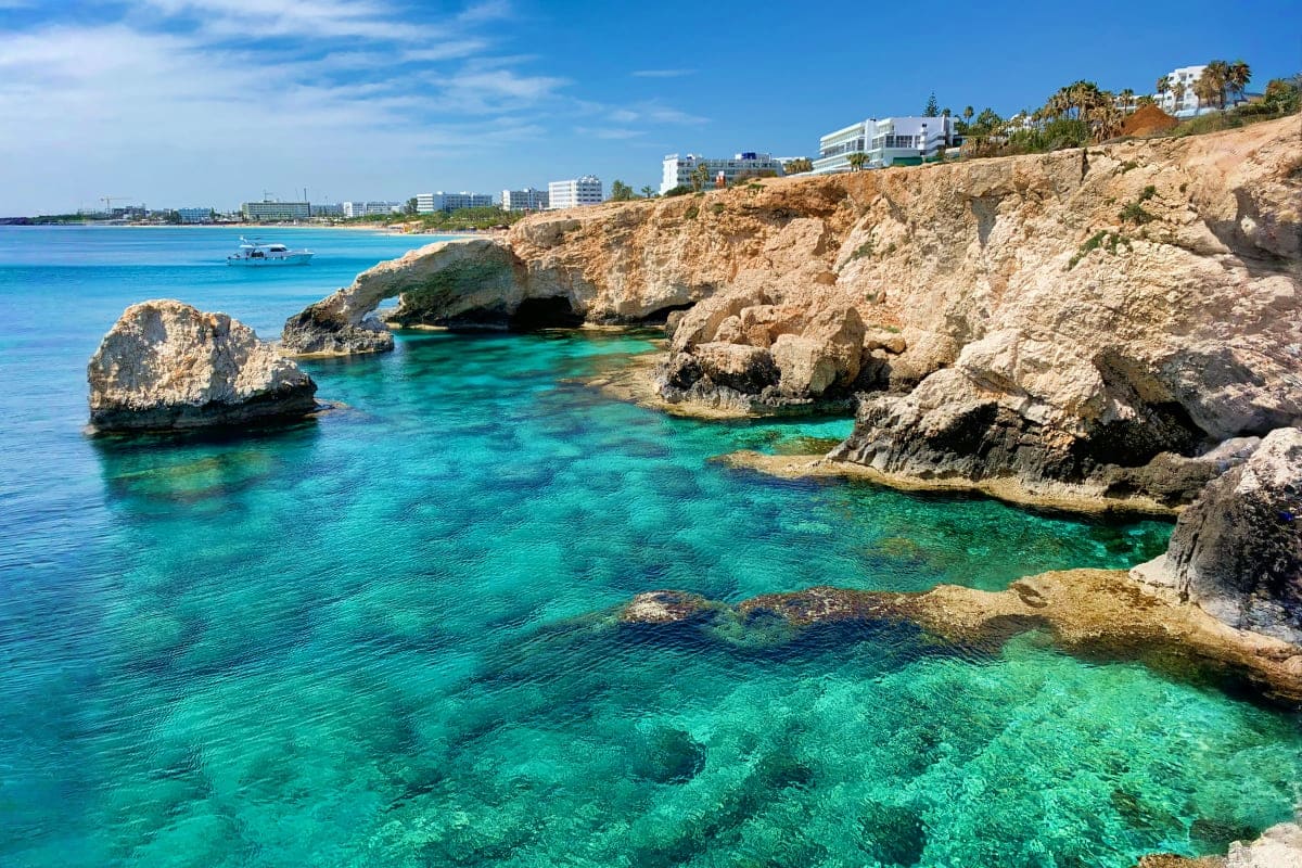 Turquoise Mediterranean waters and limestone cliffs with rock formations along a Cyprus coastal cove, hotels visible on hills