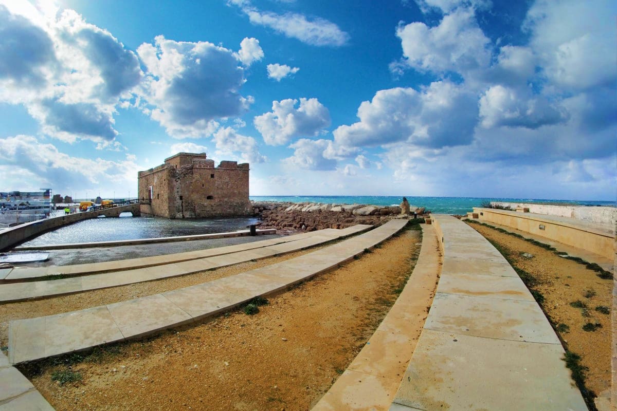 Historic stone fortress tower overlooking Mediterranean Sea with sandy beach, concrete pathways, and blue sky with white clou