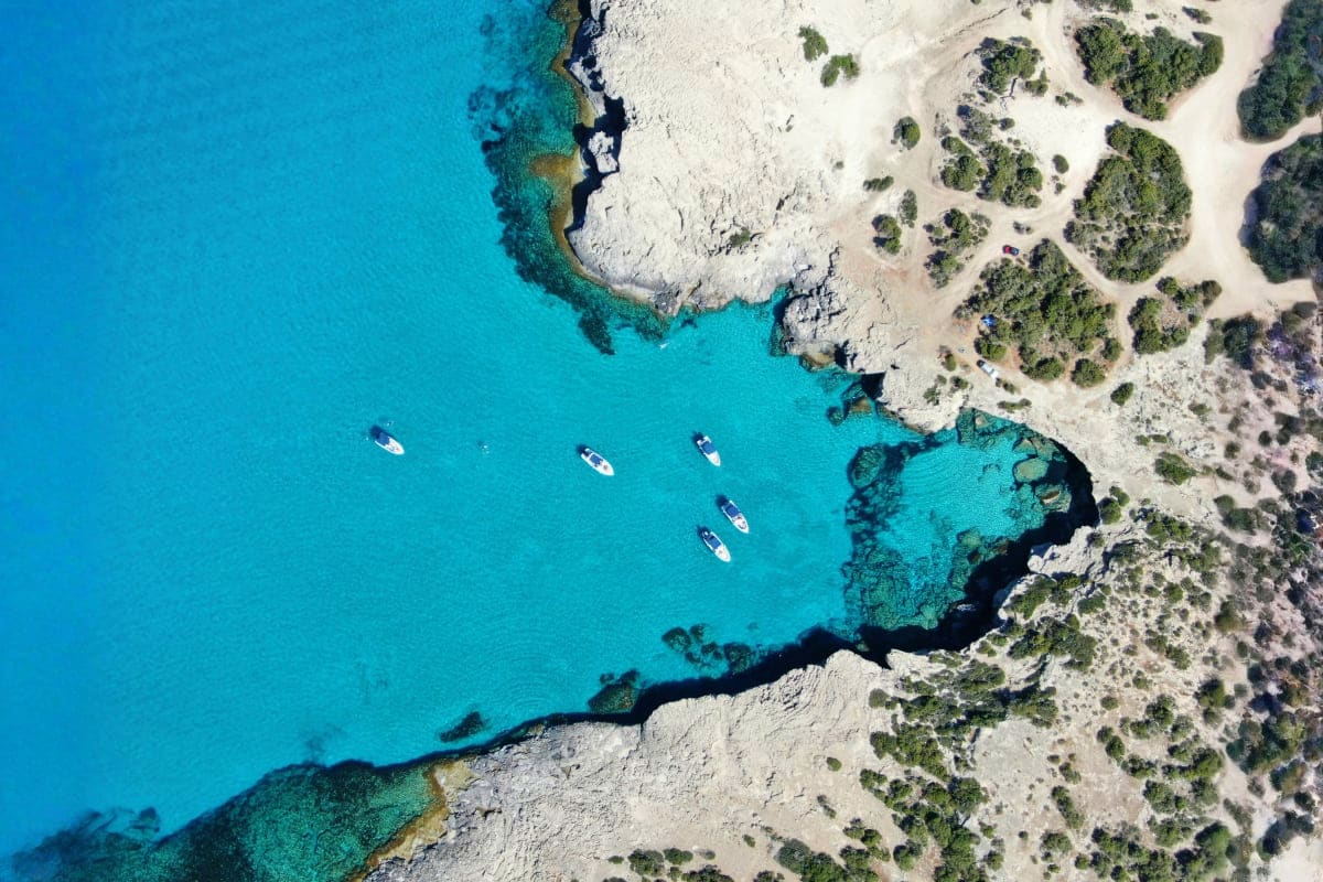 Aerial view of turquoise Mediterranean waters with white rocky cliffs, anchored boats, and green vegetation along a Cyprus co