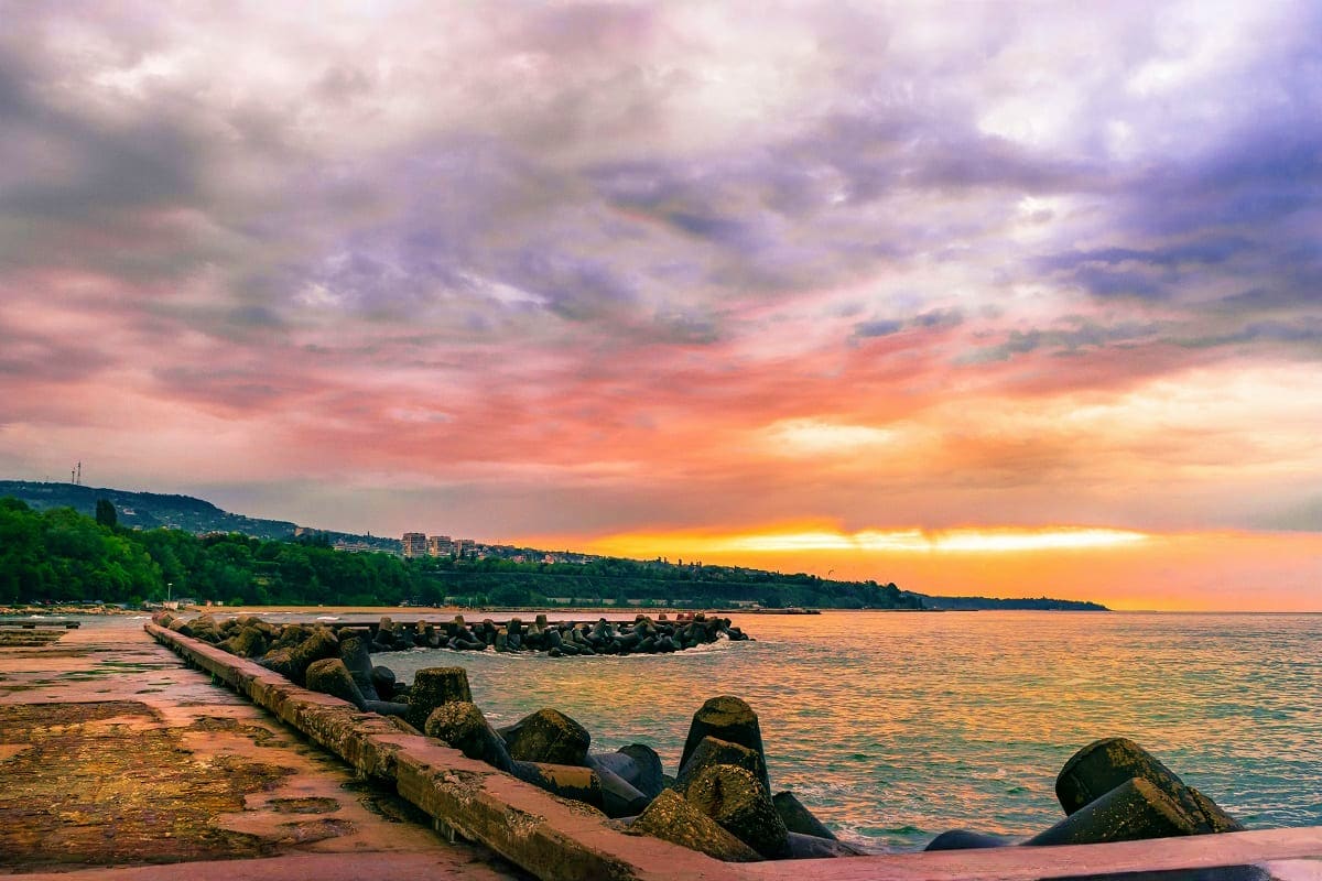Coastal sunset over Black Sea with rocky breakwater, sandy beach, and hillside town silhouette at dusk