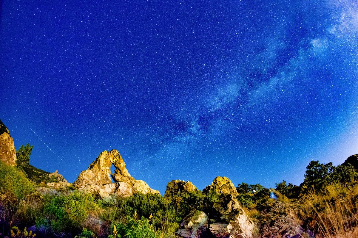 Milky Way galaxy stretching across deep blue night sky above rocky limestone formations and green vegetation in Bulgarian cou