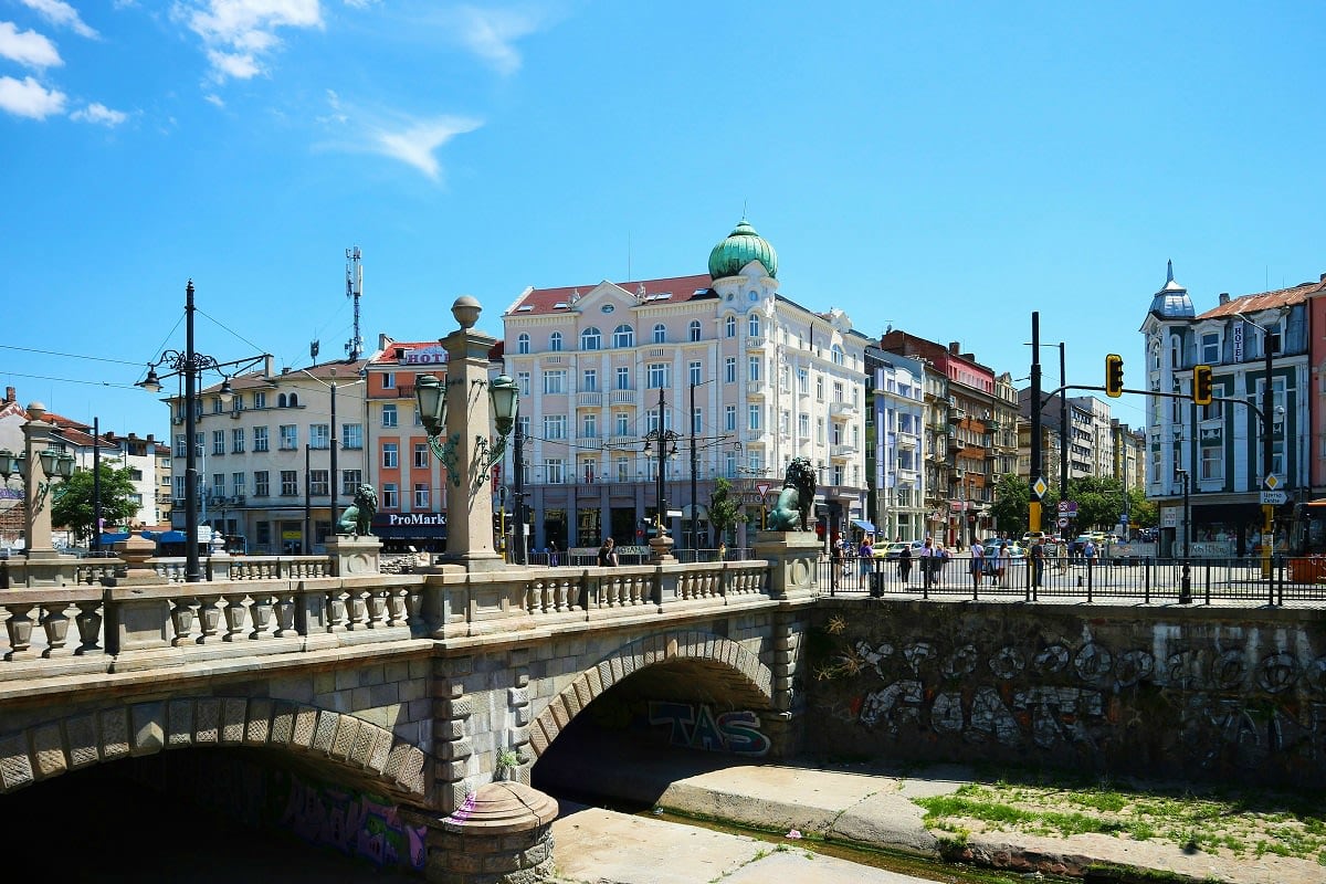 Stone bridge with statue over river in historic European city square with classical buildings and green dome architecture