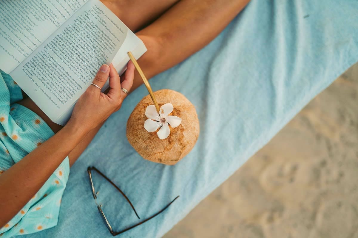 Person reading book on beach holding coconut drink with white flower garnish and sunglasses nearby