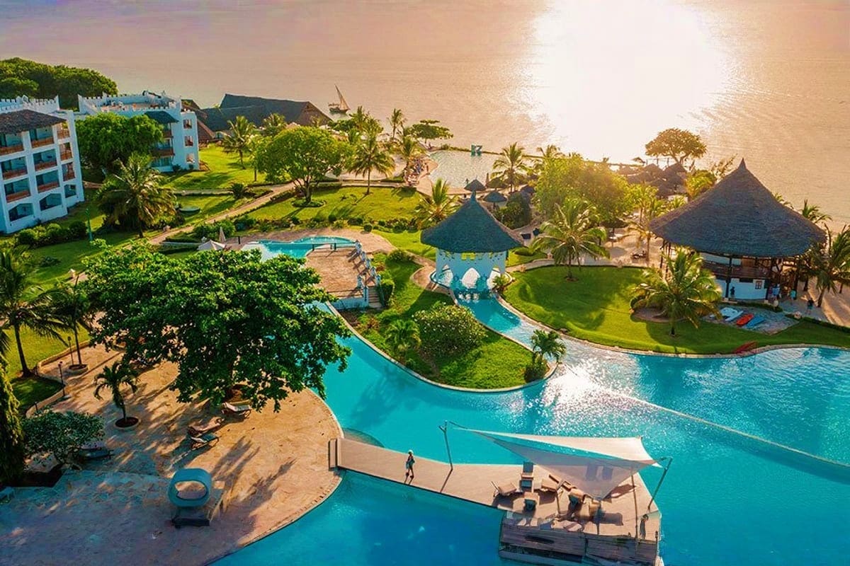 Aerial view of beachfront resort with turquoise pools, thatched-roof structures, palm trees, sandy beach, and ocean at sunset