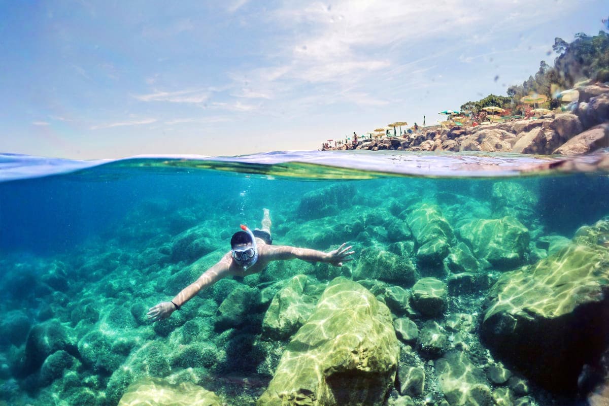 Snorkeler exploring rocky underwater reef with clear turquoise water, rocky coastline with beaches and buildings visible abov