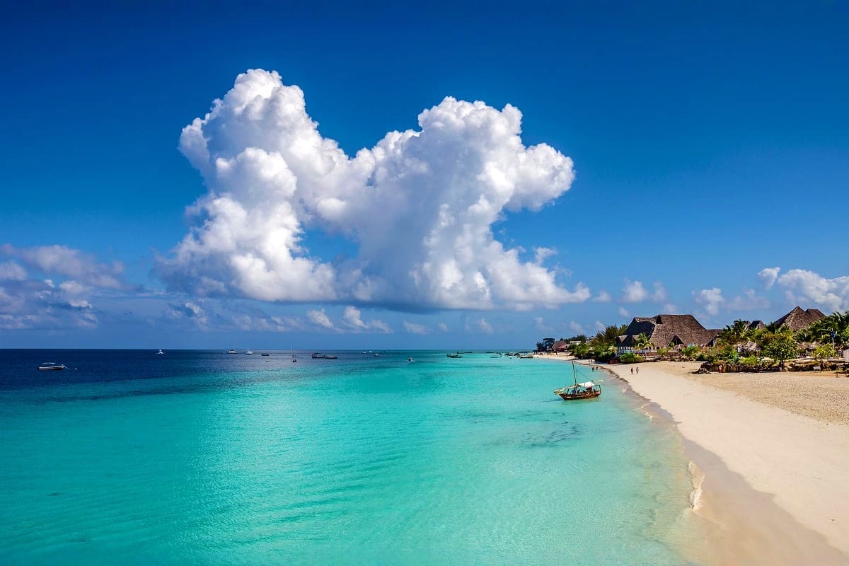 Turquoise waters and white sand beach in Zanzibar with traditional boats, thatched-roof buildings, and dramatic white clouds 