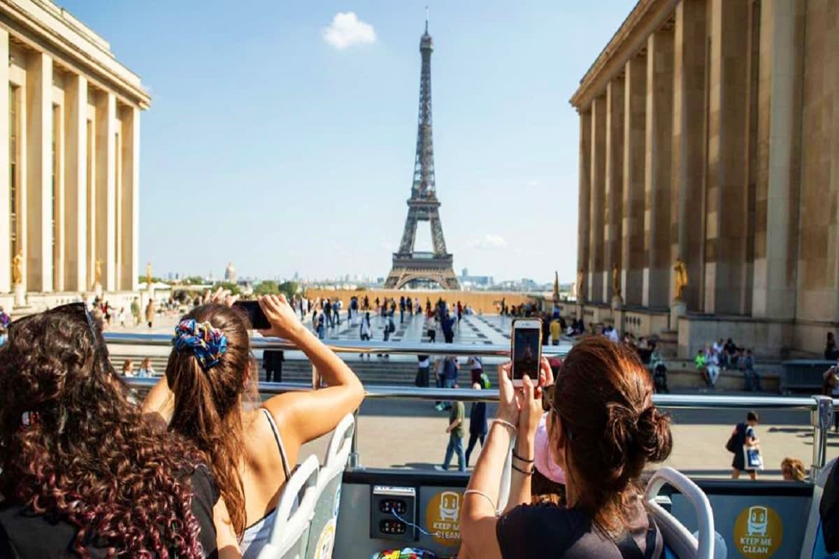 Tourists photographing the Eiffel Tower from Trocadéro plaza in Paris, with classical stone buildings framing the view
