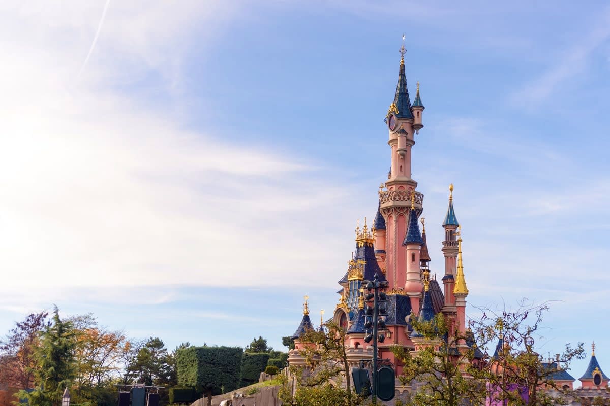 Fairy tale castle with pink and blue towers against clear sky, surrounded by autumn trees and gardens