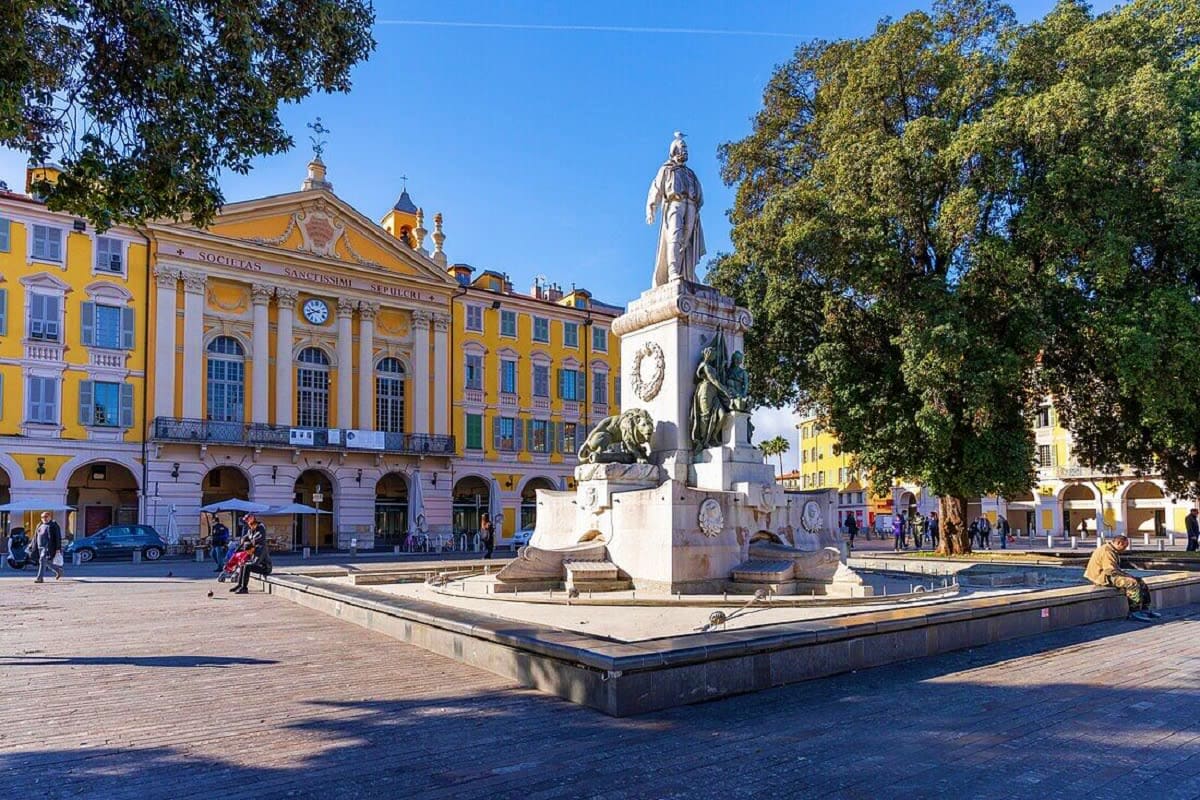 Historic square in Nice, France with white monument statue, yellow classical buildings, and trees under clear blue sky