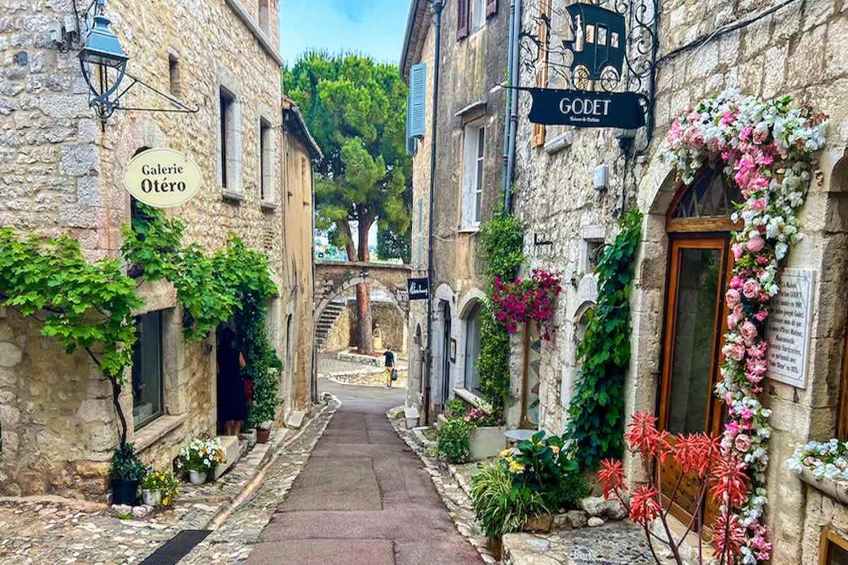 Charming cobblestone street in a medieval French village with stone buildings, flowering vines, and shop signs including Gale