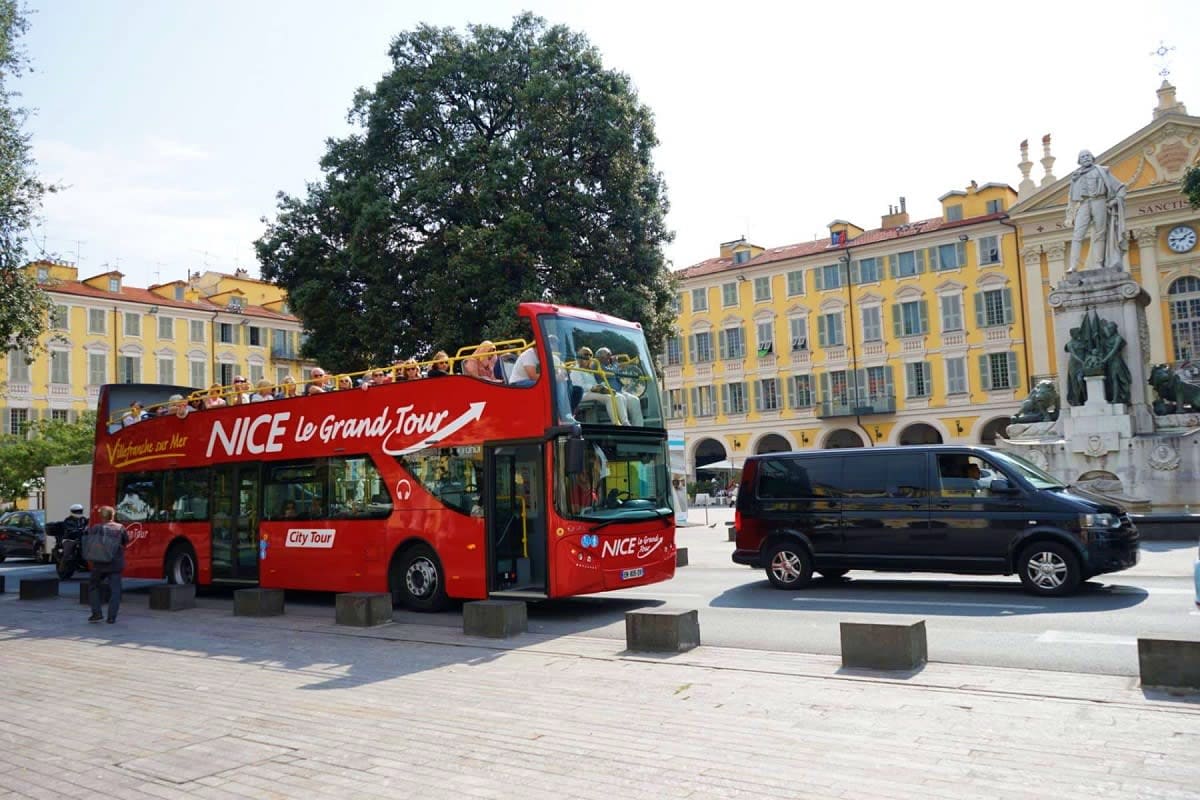 Red double-decker tour bus in Nice city square with yellow historic buildings and statue in background