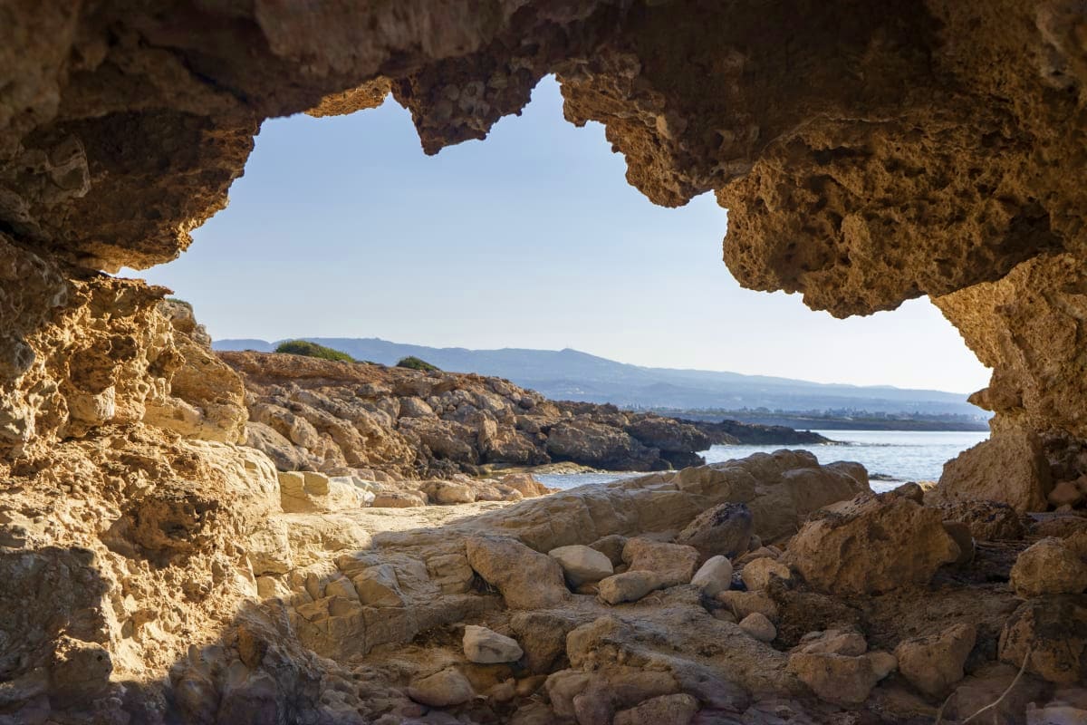 View through a limestone cave opening onto a rocky Mediterranean coastline with turquoise water and distant mountains in Cypr