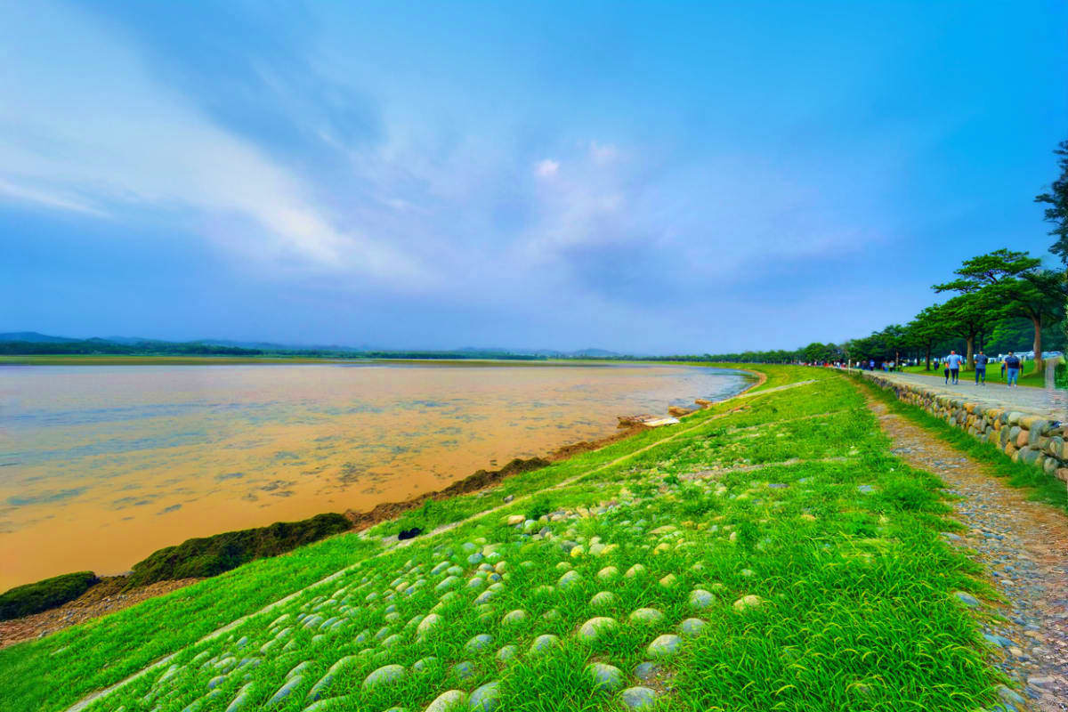 Scenic riverside embankment with lush green grass, stone pathway, and muddy river water under blue sky with distant mountains