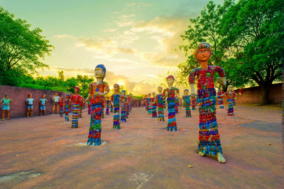 Colorful painted wooden figures in traditional dress standing in a village courtyard at sunset with trees and spectators in b
