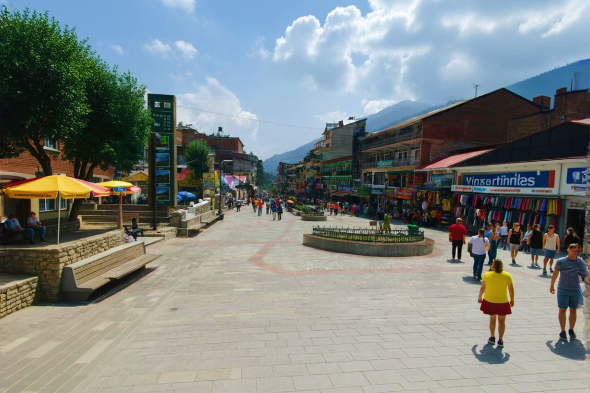Busy pedestrian plaza in Shimla with brick buildings, shops, and mountains in background under blue sky with clouds