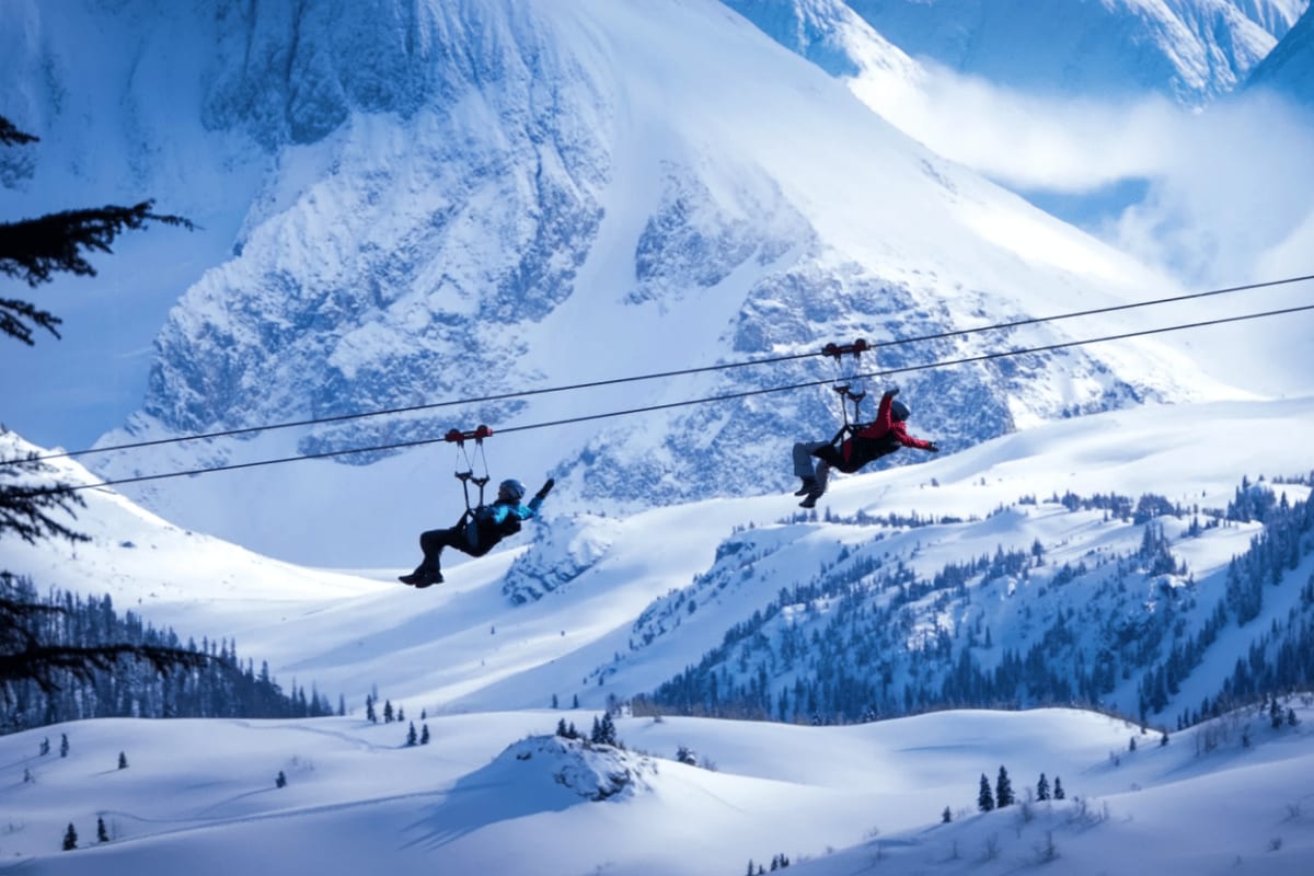 Two people zip-lining over snow-covered mountain landscape with snow-peaked mountains and evergreen forests in background