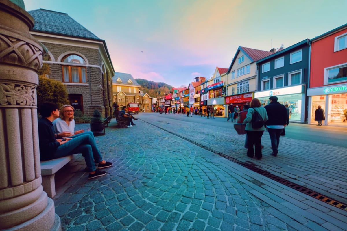 Pedestrians strolling through a colorful Alpine-style mountain town street at dusk with illuminated storefronts and snow-capp