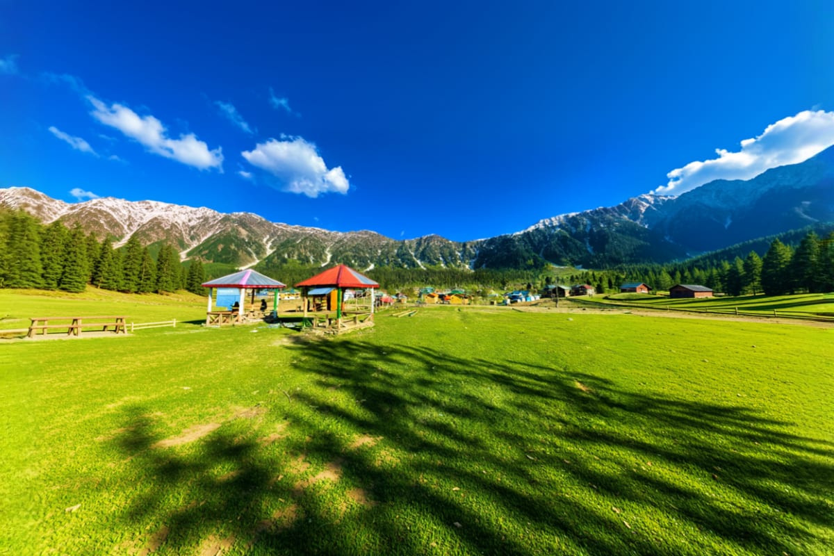 Colorful play structures with red roofs in green meadow surrounded by snow-capped mountains and forested valleys in Himachal 